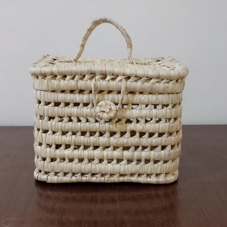 May include: A small, rectangular woven basket with a handle and a button closure. The basket is made of natural straw and has a light beige color.