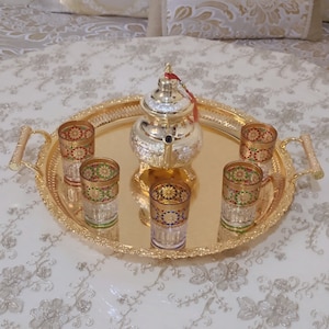May include: A gold serving tray with ornate detailing holds a silver teapot and six small glasses with colorful designs. The glasses are filled with a clear liquid.
