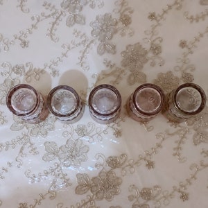 May include: Five clear glass shot glasses with a pink tint and a decorative pattern around the rim. The glasses are arranged in a row on a white surface with a floral pattern.
