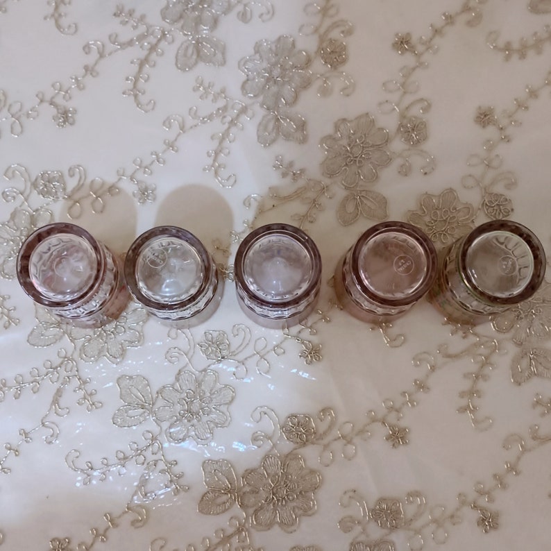 May include: Five clear glass shot glasses with a pink tint and a decorative pattern around the rim. The glasses are arranged in a row on a white surface with a floral pattern.