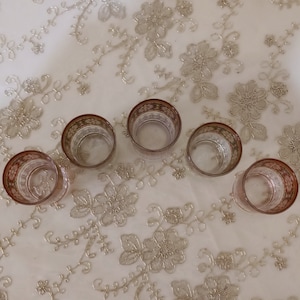 May include: Set of six clear glass shot glasses with a decorative gold and red rim. The glasses are arranged in a circle on a white tablecloth with a floral pattern.
