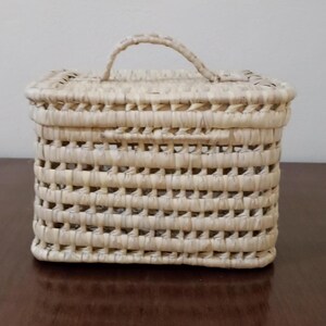 May include: A small, square, woven basket with a handle. The basket is made of natural straw and has a light beige color.
