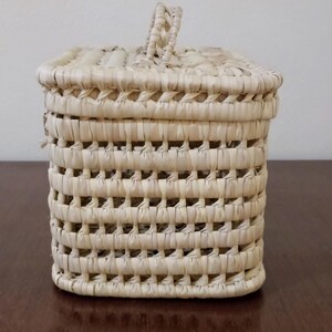 May include: A small, square, woven basket with a handle. The basket is made of natural straw and has a light beige color.