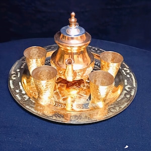 Puede incluir: Un juego de té de metal dorado con una tetera y cuatro tazas en una bandeja a juego. La tetera tiene un diseño adornado y una tapa plateada. Las tazas también están decoradas con intrincados patrones.