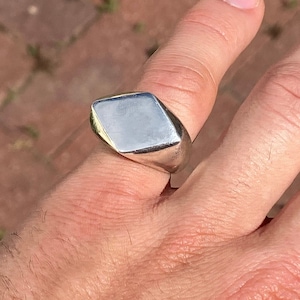 以下が含まれることがあります： 平らな長方形のトップを持つシルバーのシグネットリング。リングは指につけられています。