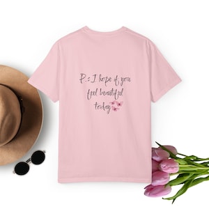 Puede incluir: Una camiseta de manga corta de color rosa claro con el texto impreso en negro P.s I hope if you feel beautiful today y tres pequeñas ilustraciones de mariposas rosas. La camiseta se muestra con un sombrero beige, gafas negras y tulipanes rosas.