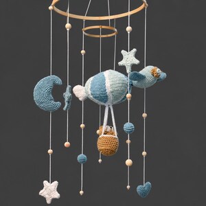 May include: A hanging mobile featuring crocheted elements suspended from a wooden ring. The design includes a blue and white striped hot air balloon with a brown basket, a crescent moon, stars, and small spheres, all accented with wooden beads.