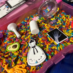 May include: A pink plastic bin filled with colorful dyed rice and small toys. The toys include a white cylinder, a magnifying glass, an avocado, a dress, and a small X-ray image. A pink scoop is in the bin, and cards with text are visible.