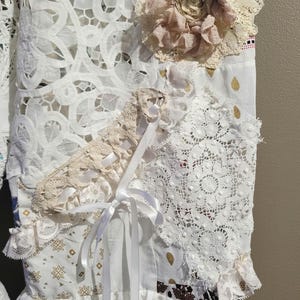 May include: A white cotton dress with lace trim and a white satin ribbon tied at the waist. The dress is decorated with various lace appliques and a large beige flower.