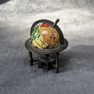 Pode incluir: Um apontador de lápis em miniatura em forma de globo com um mapa colorido com continentes amarelos, verdes e vermelhos. O globo está montado num suporte metálico escuro com uma base circular e um anel meridiano. A abertura fica na base.