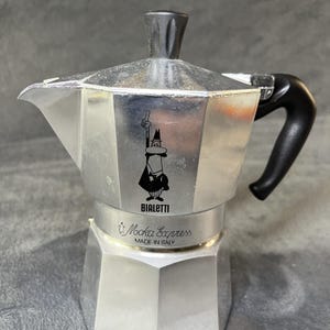 May include: A silver-coloured aluminium stovetop espresso maker featuring a black plastic handle and lid knob. The body displays the Bialetti logo with a cartoon figure and the text Moka Express MADE IN ITALY printed on the central metal band.