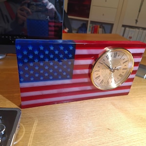 Könnte beinhalten: Eine rechteckige Tischuhr aus einem Harzblock mit dem Design der amerikanischen Flagge in Rot, Weiß und Blau. Das Zifferblatt ist kreisförmig mit einem goldfarbenen Metallrand, schwarzen Zeigern und römischen Ziffern auf der rechten Seite.