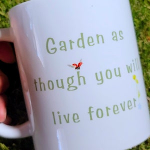 A white mug with the words "Garden as though you will live forever" on it in green letters. There is the image of a ladybug landing on the "h"