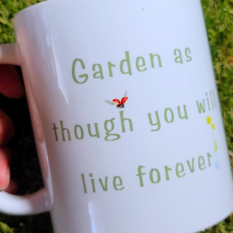 A white mug with the words "Garden as though you will live forever" on it in green letters. There is the image of a ladybug landing on the "h"