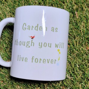 Może przedstawiać: Biały kubek ceramiczny z zielonym i czarnym wzorem. Kubek ma nadrukowany tekst "Garden as though you will live forever".
