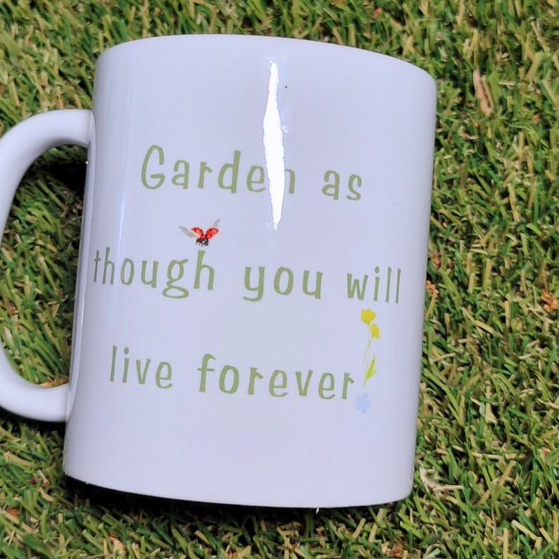 Może przedstawiać: Biały kubek ceramiczny z zielonym i czarnym wzorem. Kubek ma nadrukowany tekst "Garden as though you will live forever".