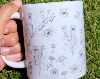 Może przedstawiać: Biały kubek ceramiczny z czarno-białym rysunkiem kwiatów nagietka i tekstem "Calendula's Garden".
