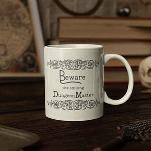 May include: White ceramic mug with a black and white design. The mug features a decorative border and the text "Beware the smiling Dungeon Master".
