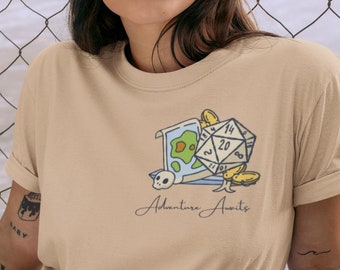 May include: A beige t-shirt with a graphic of a map, a skull, and a twenty-sided die with the text "Adventure Awaits".