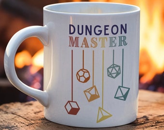 Op de afbeelding: Een witte keramische mok met de tekst "DUNGEON MASTER" in regenboogkleuren en een ontwerp van vijf kleurrijke dobbelstenen die aan touwtjes hangen. De mok staat op een houten oppervlak voor een wazige achtergrond van een kampvuur.