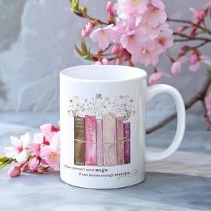 Könnte beinhalten: Weiße Keramiktasse mit einem Design aus gestapelten Büchern, aus denen Blumen wachsen. Die Tasse hat einen Henkel und den Text "One doesn't need magic if one knows enough stories..." auf der Unterseite.