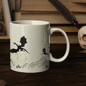 Puede incluir: Taza de cerámica blanca con una ilustración en blanco y negro de dos dragones volando sobre una cordillera. La taza tiene un asa negra y está sobre una mesa de madera.