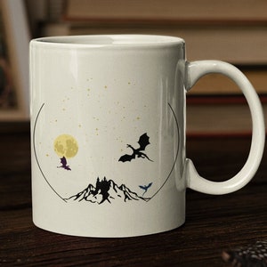 May include: White ceramic mug with a black silhouette of a mountain range and three dragons flying in front of a yellow moon. The mug has a black outline around the design.
