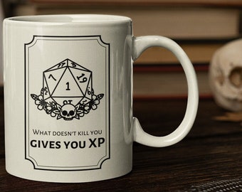 Op de afbeelding: Een witte keramische mok met een zwart-witte illustratie van een 20-zijdige dobbelsteen met het nummer 1 erop. De tekst "What doesn't kill you gives you XP" staat onder de dobbelsteen.