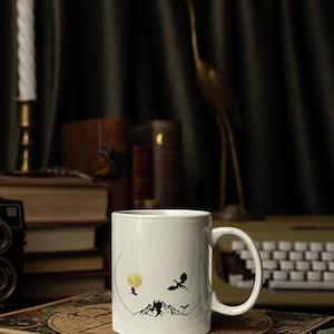 May include: White ceramic mug with a black silhouette design of a mountain range, a full moon, and three dragons flying in the sky. The mug is sitting on a book with a world map cover.