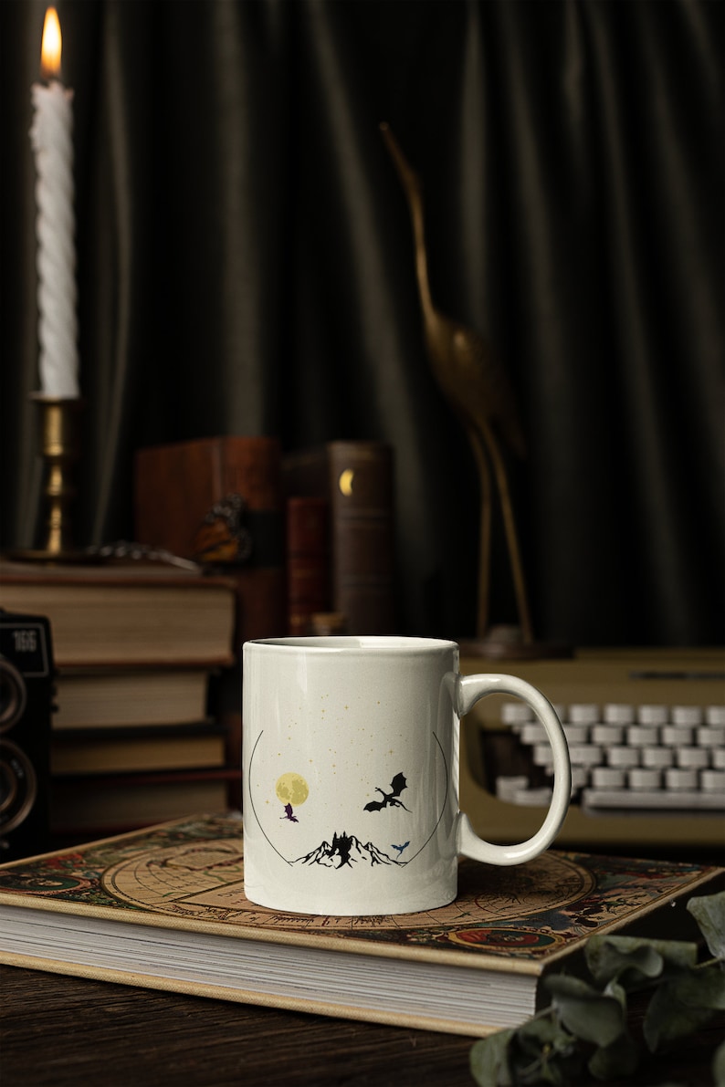 May include: White ceramic mug with a black silhouette design of a mountain range, a full moon, and three dragons flying in the sky. The mug is sitting on a book with a world map cover.