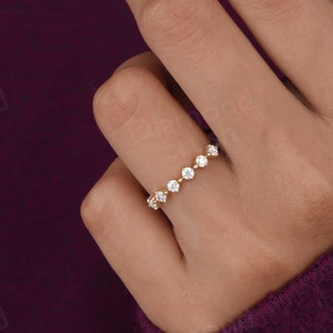 Pode incluir: Um anel de banda de ouro com uma fileira de pequenas pedras redondas transparentes. O anel está no dedo de uma pessoa.