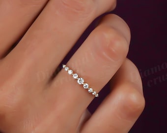 May include: A delicate gold ring with small, round, clear gemstones set along the band. The ring is shown on a hand with a burgundy sleeve in the background. The ring is a simple, elegant design.