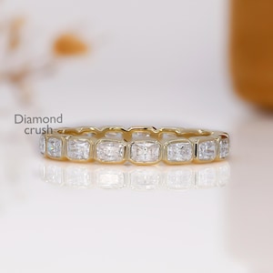May include: A gold band ring with square-cut, clear gemstones. The ring is set with multiple stones, creating a continuous pattern. The ring is on a reflective surface. The text "Diamond crush" is visible.