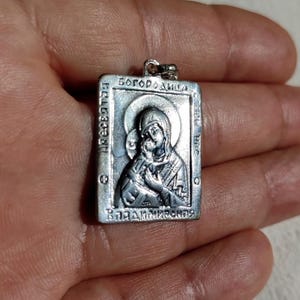 May include: Silver pendant featuring a religious icon of the Virgin Mary and baby Jesus. The icon is surrounded by a border with the text "Богородица" and "ВЛАДИМИРСКАЯ".