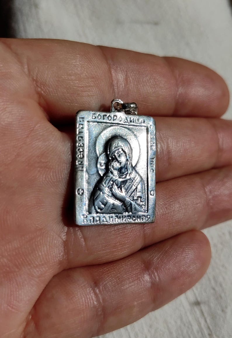 May include: Silver pendant featuring a religious icon of the Virgin Mary and baby Jesus. The icon is surrounded by a border with the text "Богородица" and "ВЛАДИМИРСКАЯ".