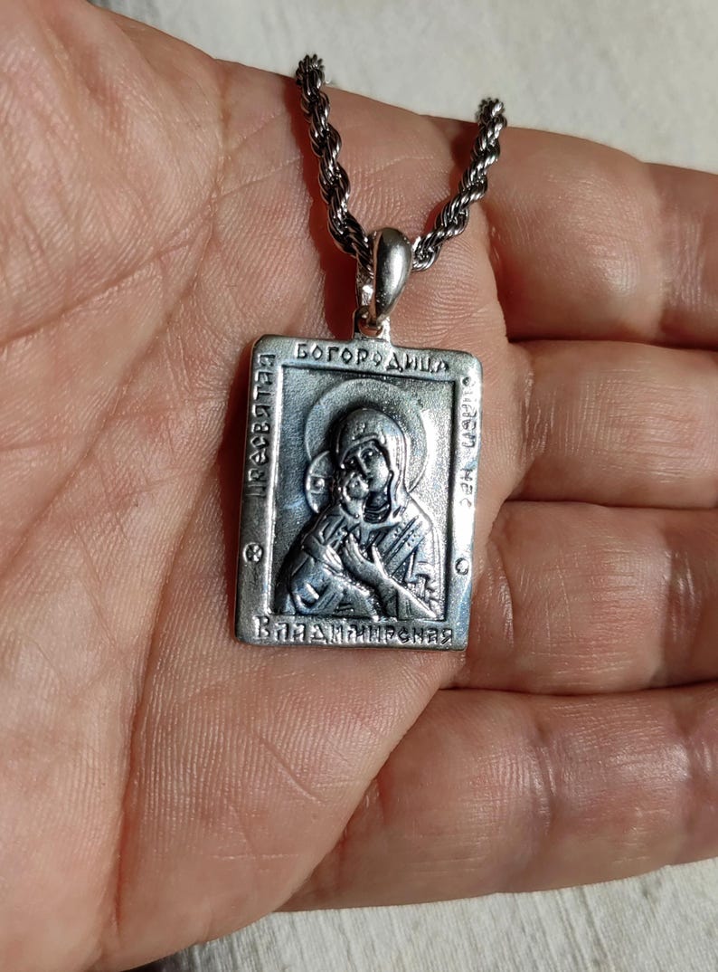 May include: Silver pendant necklace with a religious icon of the Virgin Mary and baby Jesus. The pendant is rectangular and features the inscription "Богородица" in Cyrillic script.