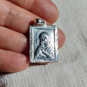 May include: Silver pendant with a religious icon of the Virgin Mary and baby Jesus. The pendant is rectangular and has the words "Богородица" and "Вратарница" inscribed on it.