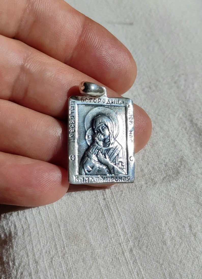 May include: Silver pendant with a religious icon of the Virgin Mary and baby Jesus. The pendant is rectangular and has the words "Богородица" and "Вратарница" inscribed on it.