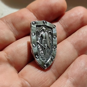 Könnte beinhalten: Silberner Schildanhänger mit einem detaillierten Bild eines Engels, der ein Schwert und einen Schild hält. Der Schild hat eine strukturierte Oberfläche und einen dekorativen Rand.