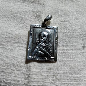 May include: Silver pendant with a religious icon of the Virgin Mary and baby Jesus. The pendant is rectangular and has a loop at the top for hanging. The icon is embossed and has a detailed design.