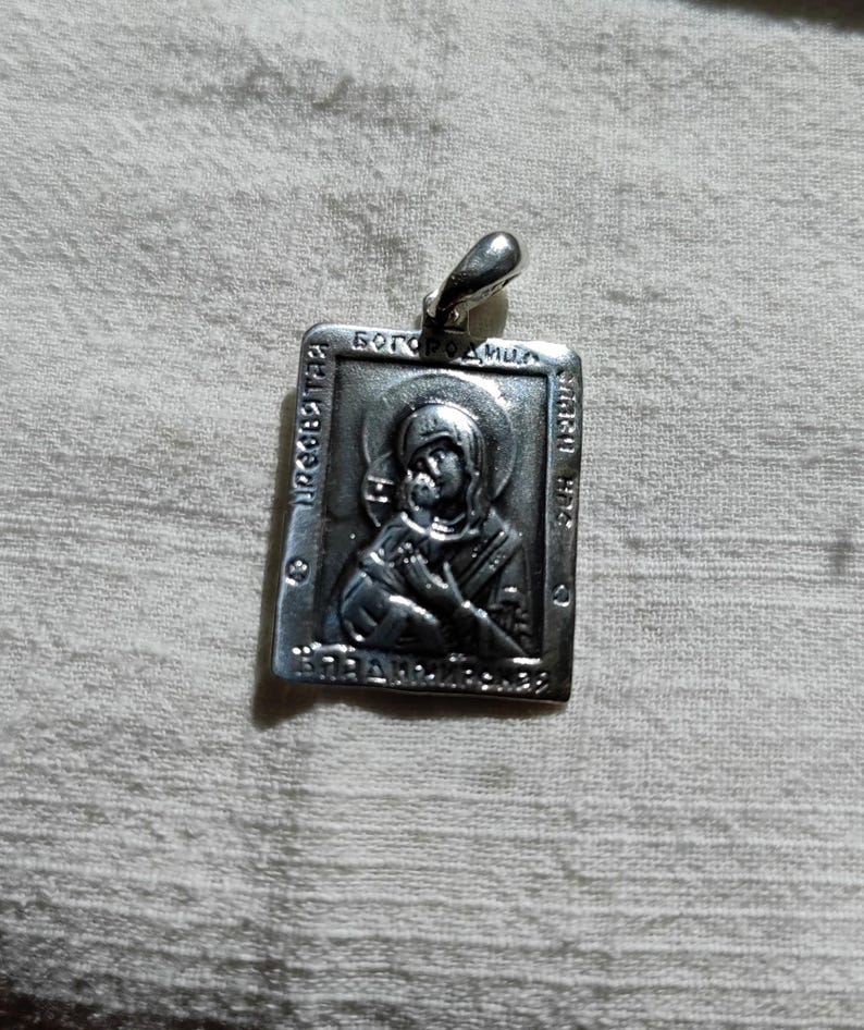 May include: Silver pendant with a religious icon of the Virgin Mary and baby Jesus. The pendant is rectangular and has a loop at the top for hanging. The icon is embossed and has a detailed design.