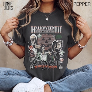 Op de afbeelding: Een antracietgrijs t-shirt met korte mouwen en een grafische print van de film Halloween III: Season of the Witch. Het ontwerp bevat een grote pompoen, personageportretten, de tekst Halloween III en Japanse karakters.