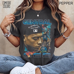 以下が含まれることがあります： 青い文字で「CENTRAL」と「CEE」と書かれたグラフィックプリントが施されたチャコールグレーのTシャツ。デザインには、野球帽をかぶった人物の大きなポートレート、座っている小さな人物、そして青と赤の様々な抽象的な要素が含まれています。