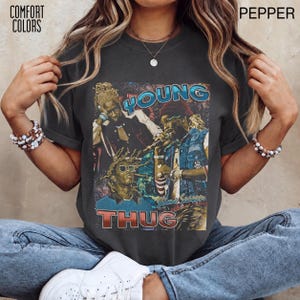 Puede incluir: Una camiseta de manga corta gris carbón con un estampado gráfico de Young Thug. El diseño incluye el texto YOUNG THUG en letras negritas, imágenes superpuestas del artista y una estética desgastada. La camiseta está etiquetada como Comfort Colors Pepper.