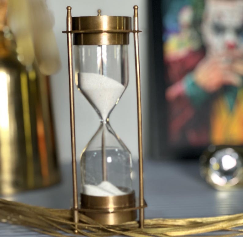 May include: A brass hourglass with white sand. The hourglass is on a table with a blurred background.