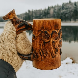 May include: A wooden mug with a carved animal handle and animal relief carvings. The mug is held by a person wearing a beige knitted glove. The mug is made of light brown wood. The mug's design suggests a rustic, outdoorsy theme.
