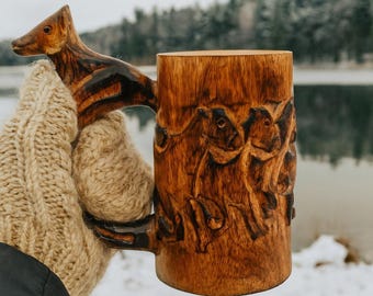 Könnte beinhalten: Ein handgefertigter Holzbecher mit einem geschnitzten Tiergriff und Reliefschnitzereien von Tieren am Körper. Der Becher wird von einer Person gehalten, die einen beigefarbenen Strickhandschuh trägt. Der Becher ist aus hellbraunem Holz.