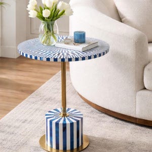 Peut inclure: Une table d'appoint ronde avec un dessus et une base à motifs bleus et blancs. La table a une tige et une base dorées. Un vase de fleurs blanches, une bougie bleue et un livre reposent sur la table. La table est à côté d'un fauteuil blanc.
