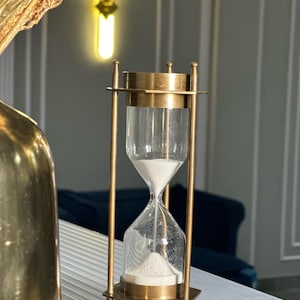 May include: A brass hourglass with a clear glass bulb. The hourglass is filled with white sand and is sitting on a white surface.