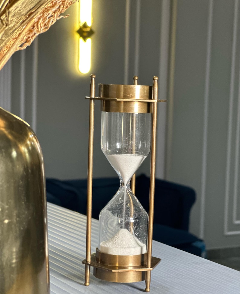 May include: A brass hourglass with a clear glass bulb. The hourglass is filled with white sand and is sitting on a white surface.